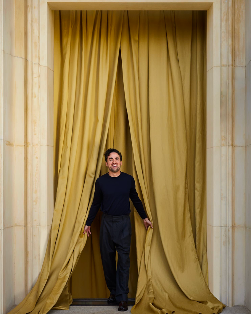 Jake Arnold standing between gold drapery panels.